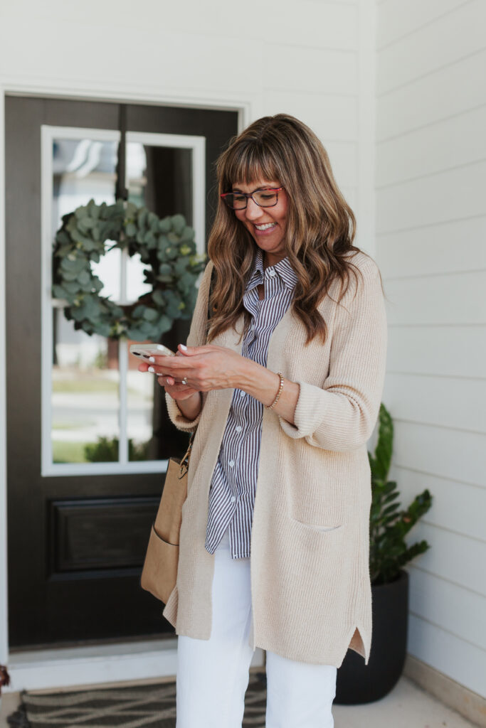 Woman smiling while checking her phone and posting to social media outside her front door, dressed in neutral, professional attire
