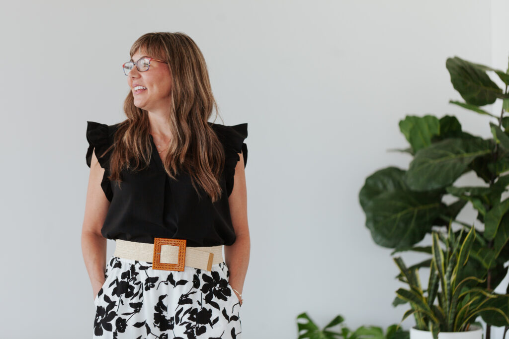 Woman in black top and patterned skirt standing indoors, smiling and looking contemplatively off-camera beside a large plant