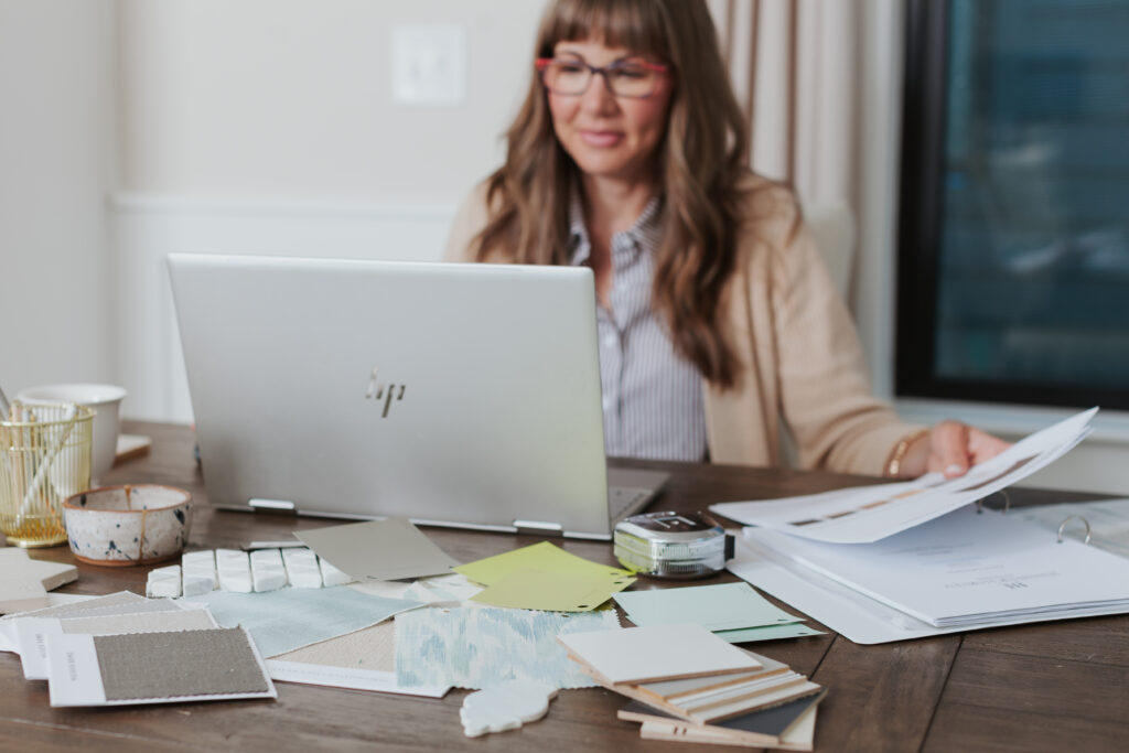 Brand photo of an interior  designer reviewing design samples and paperwork at her desk while working on a laptop in a home studio setting